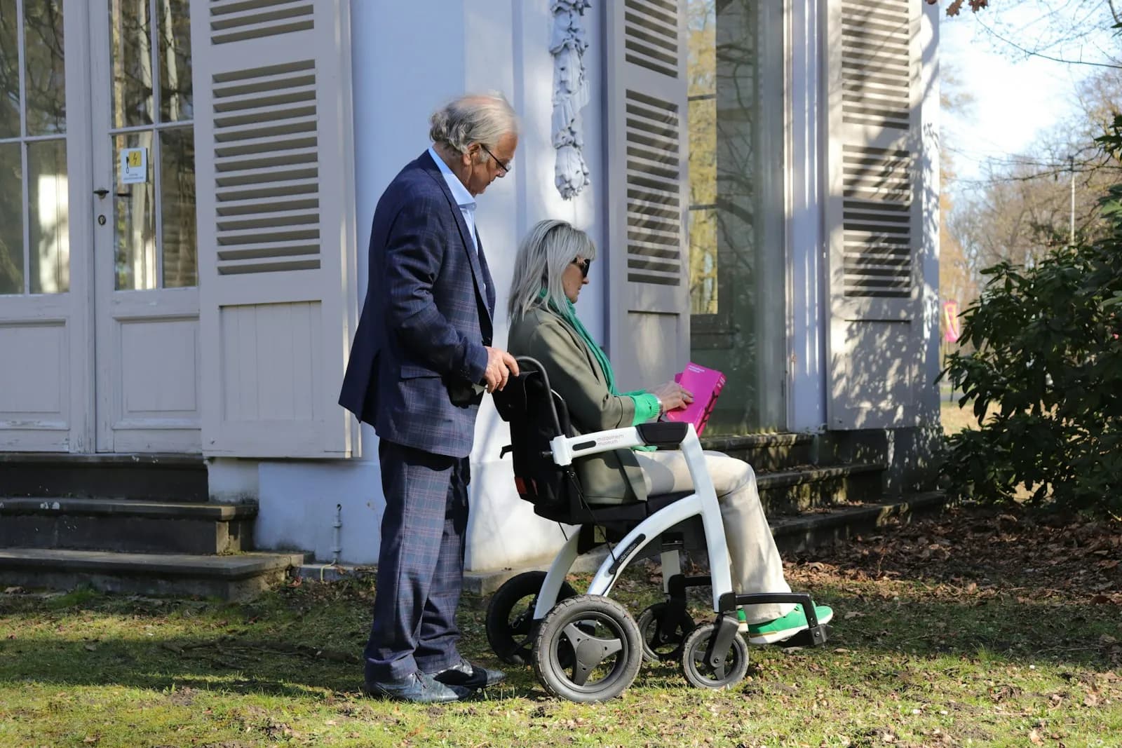 A family member pushing a relative in a wheelchair outside — the everyday physical caring that continues at home but becomes the moment Carer's Allowance and disability benefits start their 28-day countdown when the cared-for person moves into a publicly-funded care home.