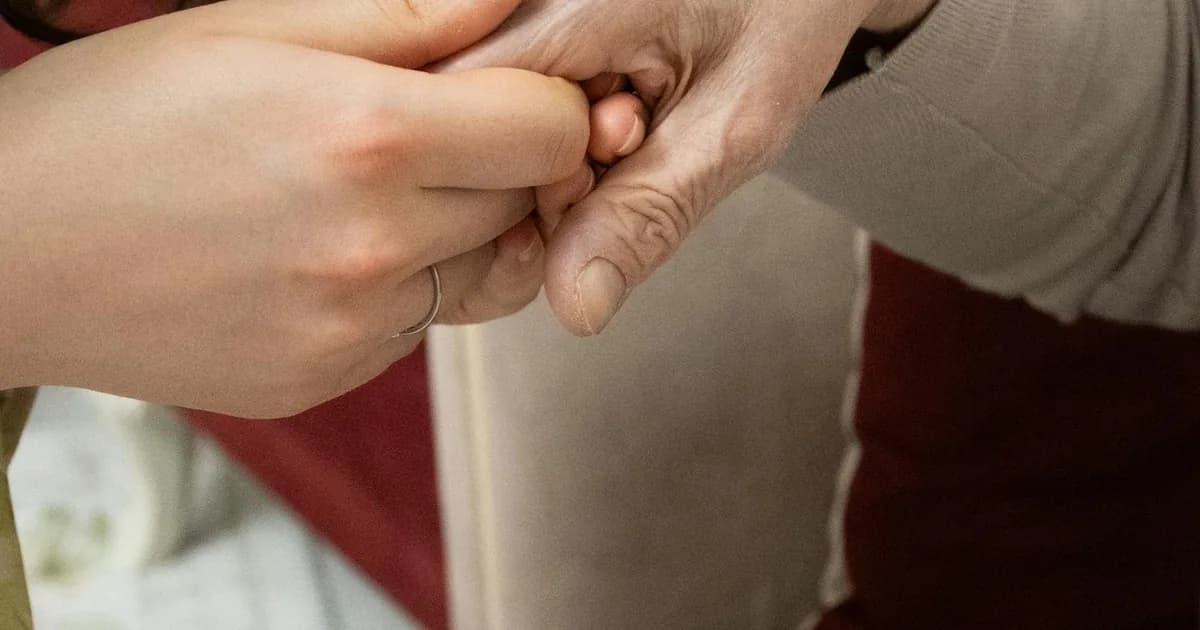 An adult child holding the hand of an older relative in a quiet care home setting, representing the lived experience of families navigating NHS Continuing Healthcare refusals.