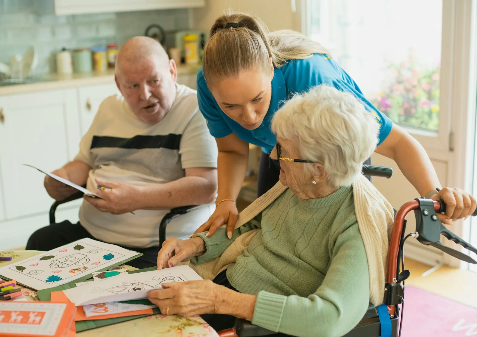 A caregiver sitting at a table with an older couple, working through a quiet activity together — the everyday relational caring at the heart of UK domiciliary care.