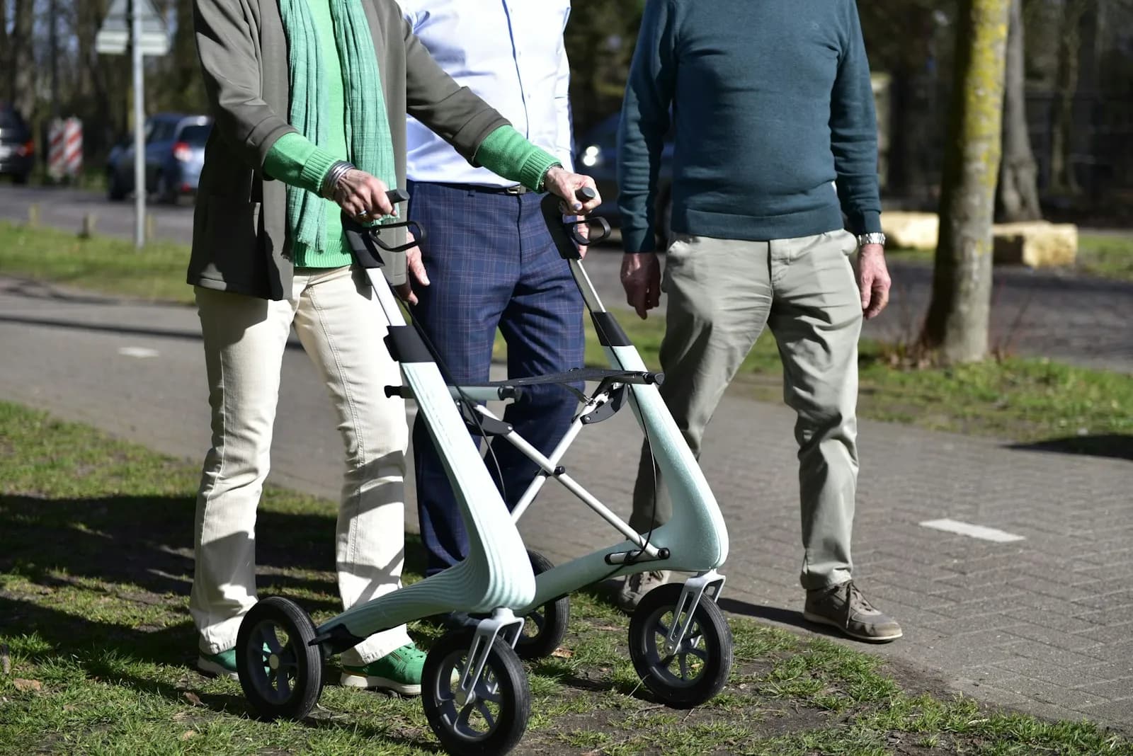 Three people walking together outdoors with a wheeled walker — the kind of mobility and falls-risk supervision that often qualifies for higher levels of domiciliary care and, in many cases, for NHS Continuing Healthcare funding.