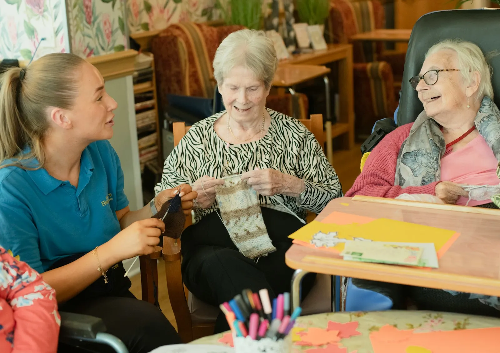 A caregiver assisting two older women with a knitting activity in a familiar living-room setting — the kind of relational, one-to-one supportive caring that defines live-in care delivery in the UK.