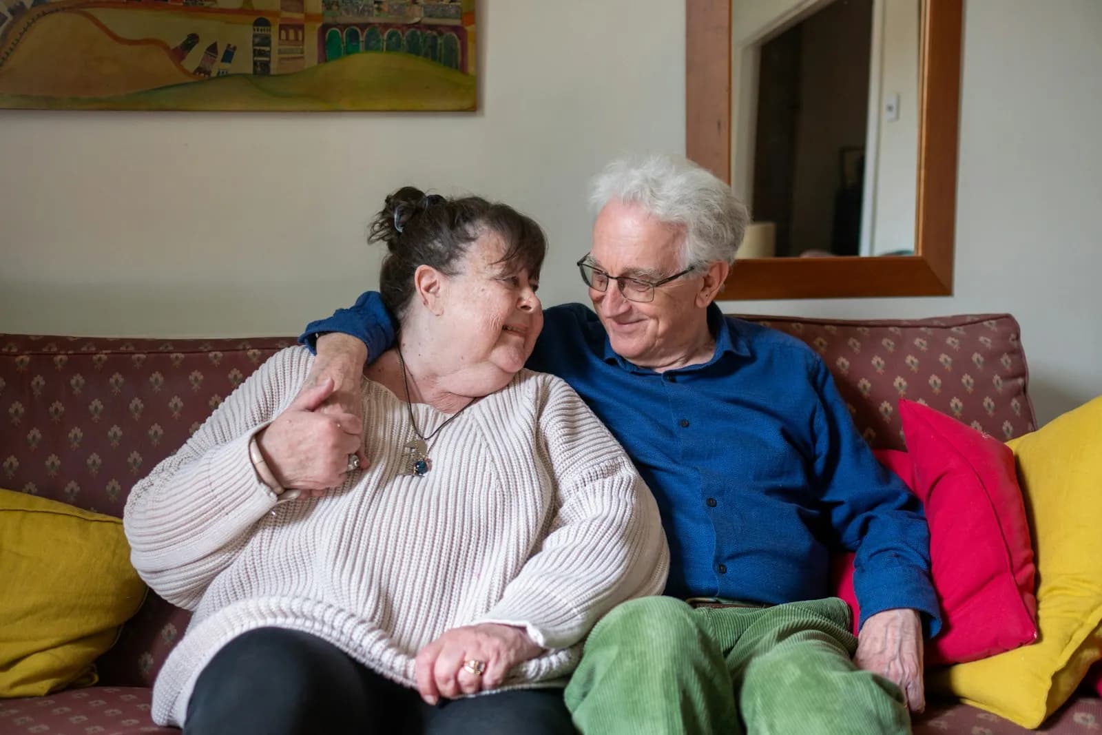 An older couple sitting together on a sofa in their familiar home setting — the cohabitation continuity that live-in care preserves for couples and the relational frame at the heart of the live-in-vs-care-home choice.