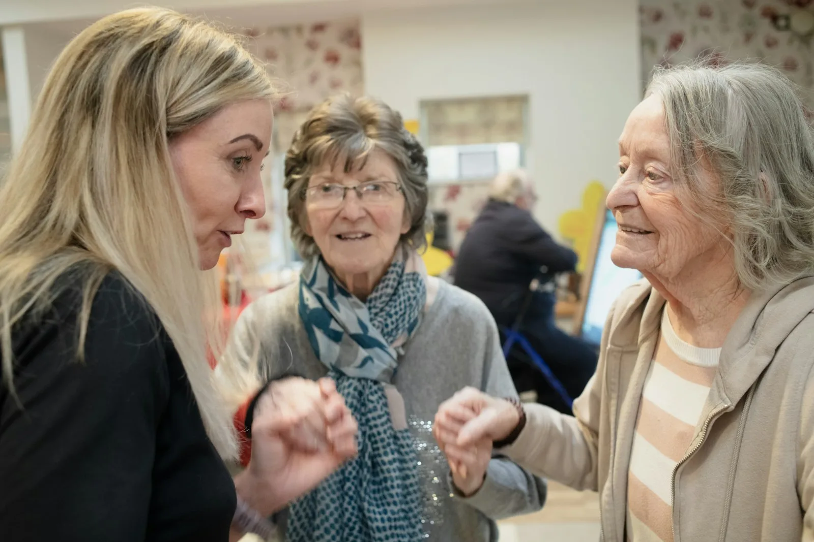 A family carer in conversation with two older women in a care home day room — the moment of arrival that respite care exists to make possible, giving the cared-for person social contact and the family carer time to recover.